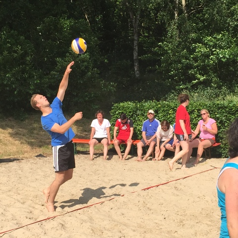 Ein Spieler mit blauem Shirt f&uuml;hrt einen Aufschlag beim Beachvolleyball aus, Zuschauer sitzen im Hintergrund.