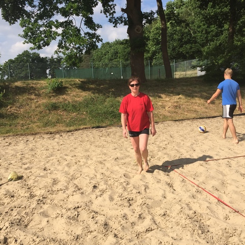 Person in rotem T-Shirt und Shorts l&auml;uft barfu&szlig; auf einem Sandplatz, im Hintergrund ein weiteren Spieler und B&auml;ume.
