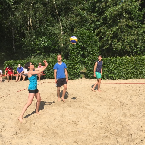 Drei Personen spielen Volleyball am Strand; eine Frau passt den Ball, w&auml;hrend zwei M&auml;nner warten.