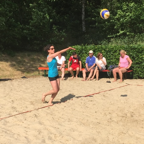 Frau in blauer Sportbekleidung spielt Beachvolleyball im Sand, Zuschauer sitzen auf einer Bank im Hintergrund.