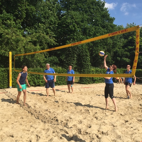 Strandvolleyballspiel mit f&uuml;nf Spielern auf Sand, Baumgr&uuml;n und blauem Himmel im Hintergrund.