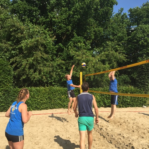 Vier Personen spielen Beachvolleyball auf einem Sandplatz, w&auml;hrend ein Spieler zum Ball springt.
