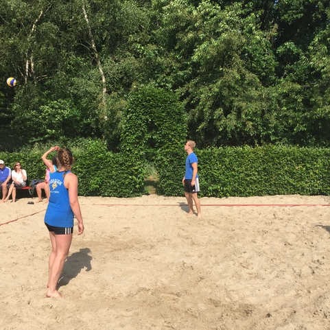 Zwei Spieler im Sandbereich werfen einen Volleyball, Zuschauer sitzen im Hintergrund auf B&auml;nken.