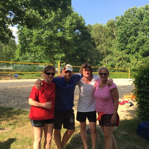 Vier Personen stehen am Sandvolleyballfeld, umgeben von gr&uuml;ner Natur, bei sonnigem Wetter.