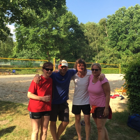 Vier Personen l&auml;cheln vor einem Volleyballfeld aus Sand, umgeben von B&auml;umen und sonnigem Wetter.