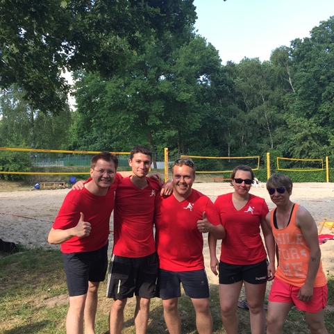 Gruppe von f&uuml;nf Personen in roten T-Shirts, l&auml;chelnd am Sandvolleyballfeld im Freien.