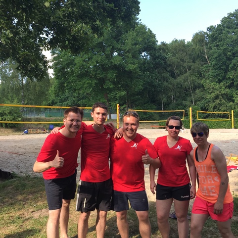 F&uuml;nf Personen in roten T-Shirts posieren l&auml;chelnd vor einem Beachvolleyballfeld mit B&auml;umen im Hintergrund.