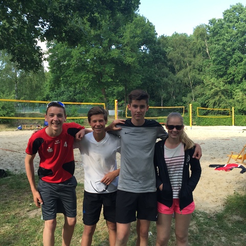 Vier Jugendliche stehen auf einer Sandfl&auml;che neben einem Volleyballfeld im Freien, umgeben von B&auml;umen.
