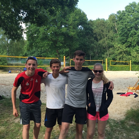Vier Jugendliche stehen l&auml;chelnd nebeneinander auf einem Sandplatz f&uuml;r Beachvolleyball, im Hintergrund Sportger&auml;te und B&auml;ume.