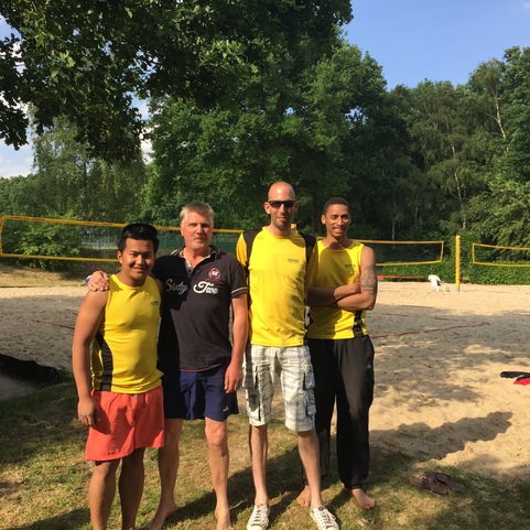 Vier M&auml;nner stehen gemeinsam am Sandvolleyballfeld, umgeben von B&auml;umen und sonnigem Wetter.