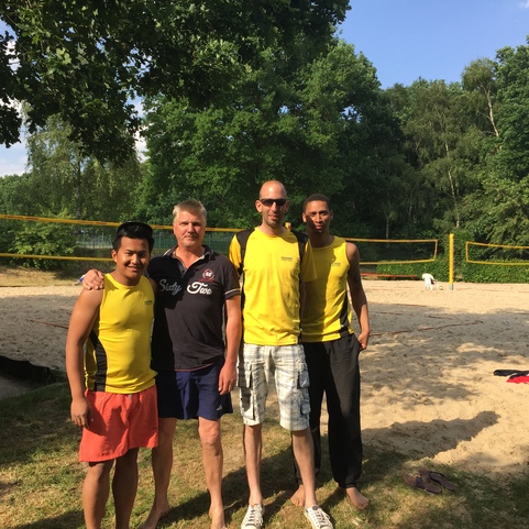 Vier M&auml;nner in gelben Tanktops stehen l&auml;chelnd vor einem Sandvolleyballfeld mit Netzen und B&auml;umen im Hintergrund.