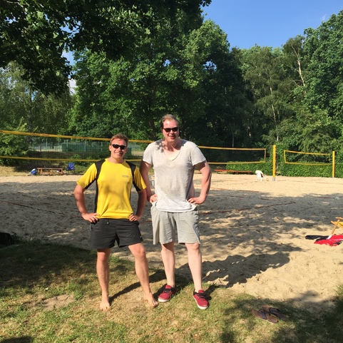 Zwei M&auml;nner stehen l&auml;ssig am Sandstrand eines Volleyballfeldes, umgeben von B&auml;umen und Sonnenschein.