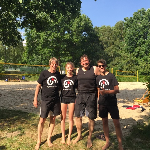 Vier Personen in Sportbekleidung stehen auf einem Sandplatz f&uuml;r Beachvolleyball, umgeben von B&auml;umen und Sonne.