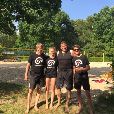 Vier Personen in Sportbekleidung stehen l&auml;chelnd am Strand eines Volleyballfeldes umgeben von B&auml;umen und Sand.