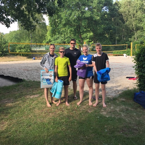 Gruppe von f&uuml;nf Personen am Sandvolleyballfeld, bekleidet in Sportkleidung, mit Handt&uuml;chern und Spielern an der Seite.