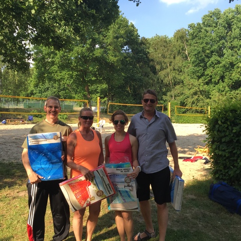 Vier Personen stehen auf einer Wiese vor einem Sandplatz mit Volleyballnetz, halten Handt&uuml;cher oder Matten.