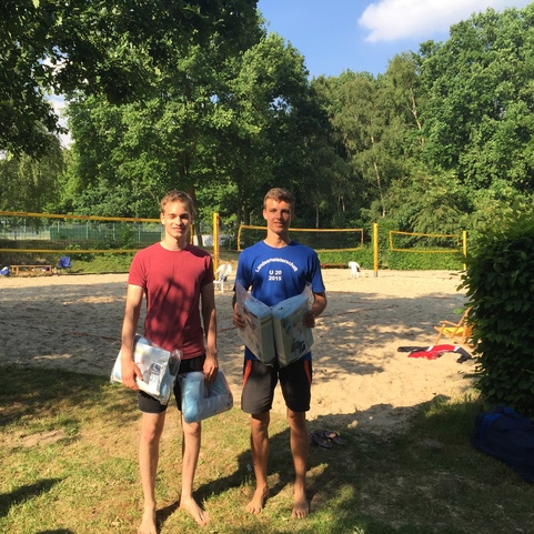 Zwei M&auml;nner stehen barfu&szlig; auf einem Sandplatz, jeder tr&auml;gt einen Volleyball und pers&ouml;nliche Gegenst&auml;nde.