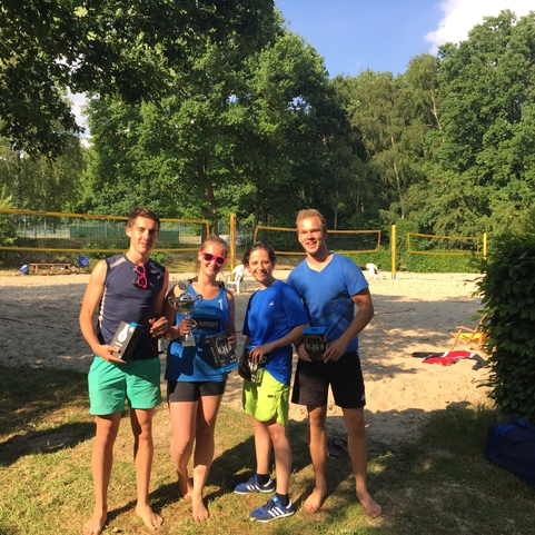 Vier Personen stehen am Strandvolleyballfeld, halten gemeinsam ihre B&auml;lle und l&auml;cheln im sonnigen Freien.