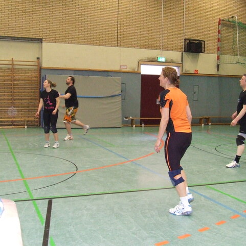 Vier Personen spielen Volleyball in einer Sporthalle mit gr&uuml;ner Fl&auml;che und Basketballnetz im Hintergrund.