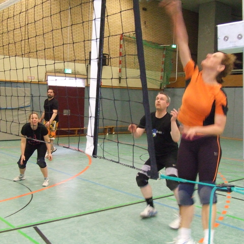 Mehrere Volleyballspieler in einer Halle w&auml;hrend eines Spiels, eine Spielerin springt, um den Ball zu treffen.