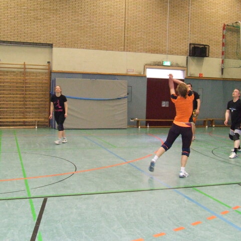 Eine Spielerin in Orange schl&auml;gt einen Volleyball auf einem Sportplatz; weitere Spielerinnen sind im Hintergrund aktiv.