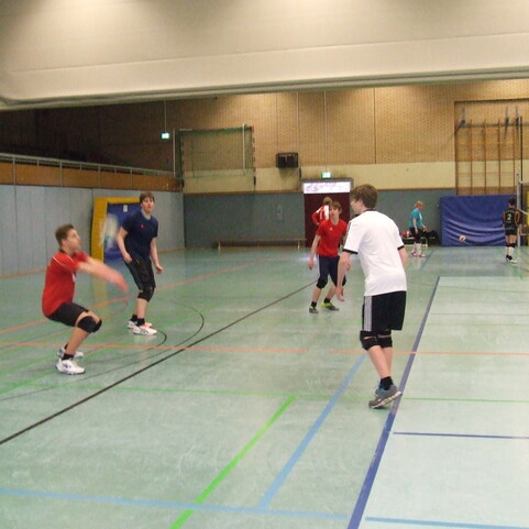Sechs junge Menschen spielen Volleyball in einer Sporthalle, mit einem Netz und Trainingsger&auml;ten im Hintergrund.