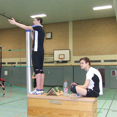 Zwei Volleyball-Schiedsrichter in einer Halle, einer steht auf einem Holzkasten, der andere sitzt daneben.