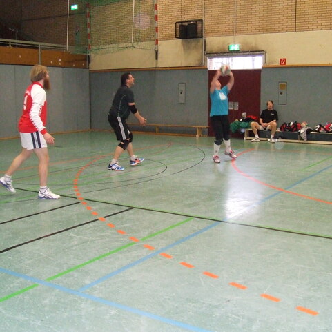 Drei Personen in Sportkleidung spielen in einer Halle Basketball. Eine Person wirft den Ball, zwei stehen bereit.