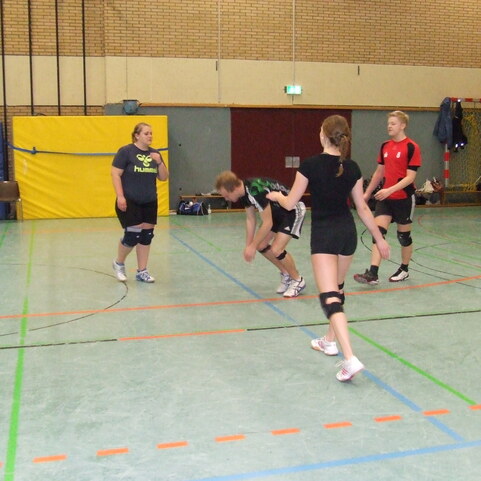 Vier Jugendliche trainieren in einer Sporthalle f&uuml;r Volleyball, mit Netzen und B&auml;llen im Hintergrund.