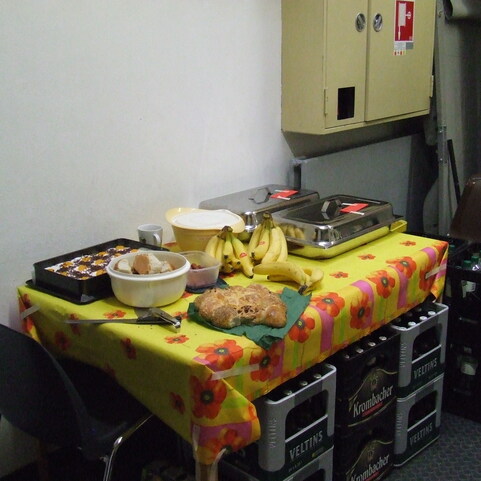Tisch mit gelber Tischdecke, darauf Bananen, Geb&auml;ck, Snacks und Getr&auml;nken, neben einem Stuhl und einer Wand.