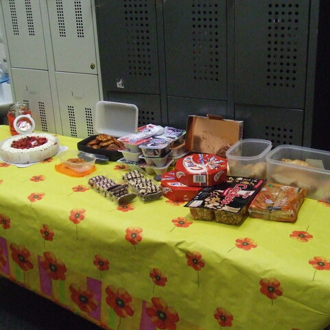 Ein Tisch mit gelber Tischdecke, belegt mit verschiedenen S&uuml;&szlig;igkeiten, Snacks und einer Torte, im Hintergrund Schlie&szlig;f&auml;cher.