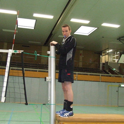 Ein Mann steht auf einer Bank neben einem Volleyballnetz und h&auml;lt einen Schiedsrichterpfeife in der Hand.