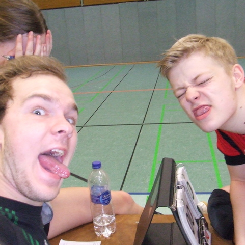 Selfie zweier junger M&auml;nner mit herausgestreckten Zungen, im Hintergrund eine Sporthalle mit gr&uuml;ner Bodenmarkierung.