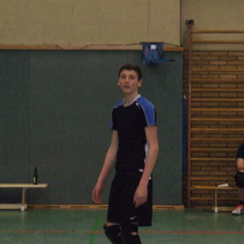 Junger Spieler in Sportkleidung steht in einer Halle, im Hintergrund sitzt ein weiterer Spieler auf einer Bank.