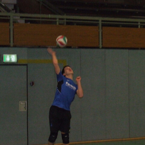 Junger Volleyballspieler in blauer Shirt, der einen Aufschlag ausf&uuml;hrt, w&auml;hrend der Ball hoch geworfen wird.