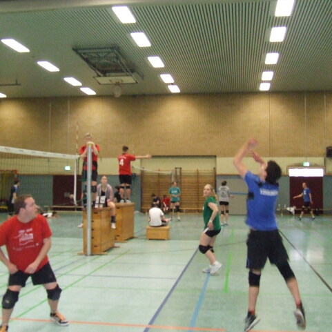 Volleyballspiel in einer Halle mit mehreren Spielern, einer springt zur Ballannahme, andere stehen in verschiedenen Positionen.