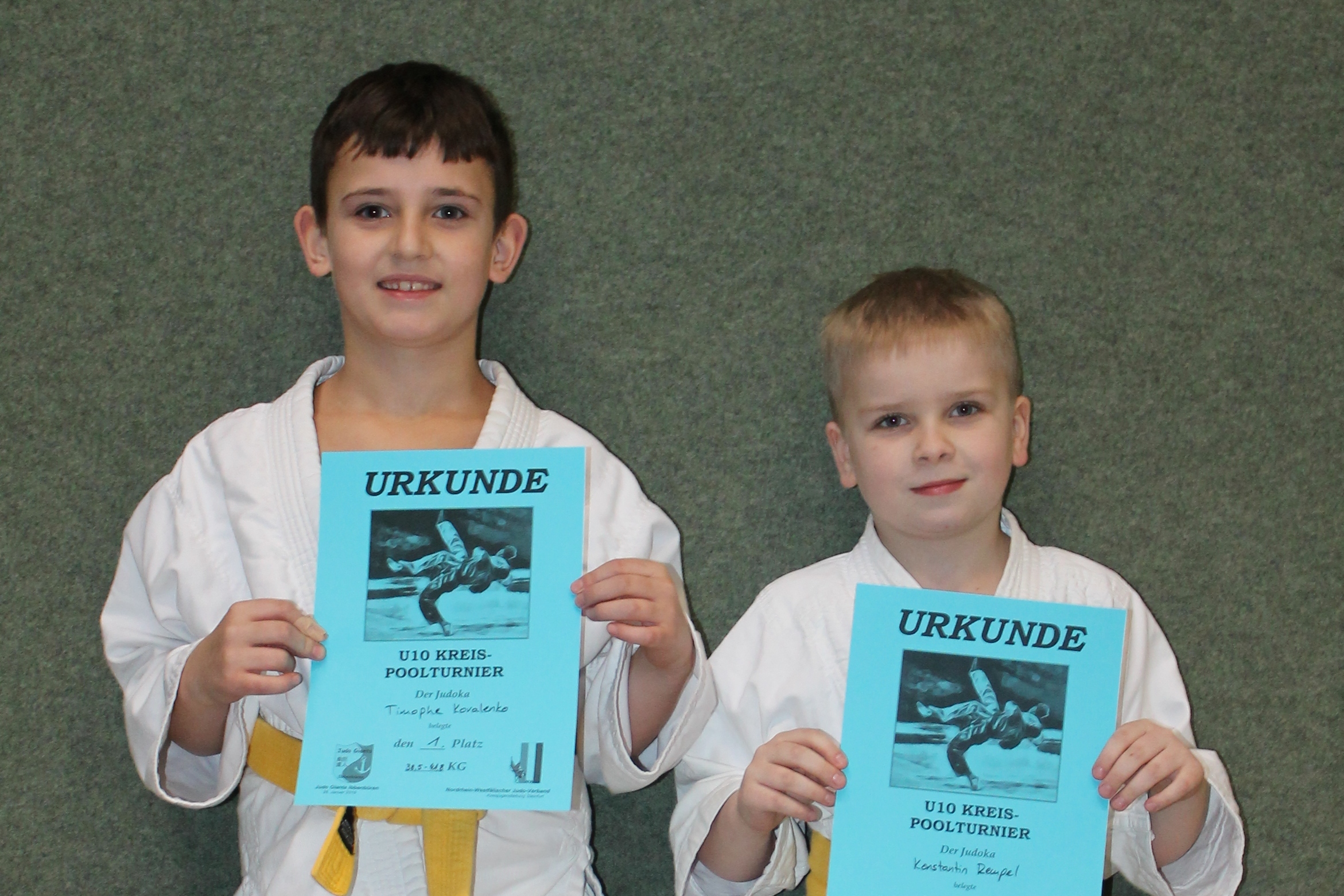 Zwei Kinder in wei&szlig;en Judoanz&uuml;gen halten stolz ihre Urkunden vor einem grauen Hintergrund.