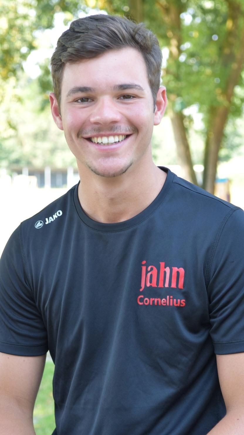 Junger Mann mit kurzem Haar, l&auml;chelt, tr&auml;gt ein schwarzes Sportshirt mit rotem Aufdruck, sitzt in einer park&auml;hnlichen Umgebung.