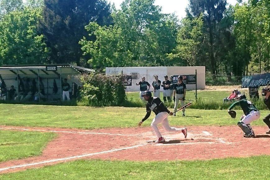 Baseballspieler schlägt einen Ball, während der Catcher in Position ist; im Hintergrund stehen weitere Spieler.