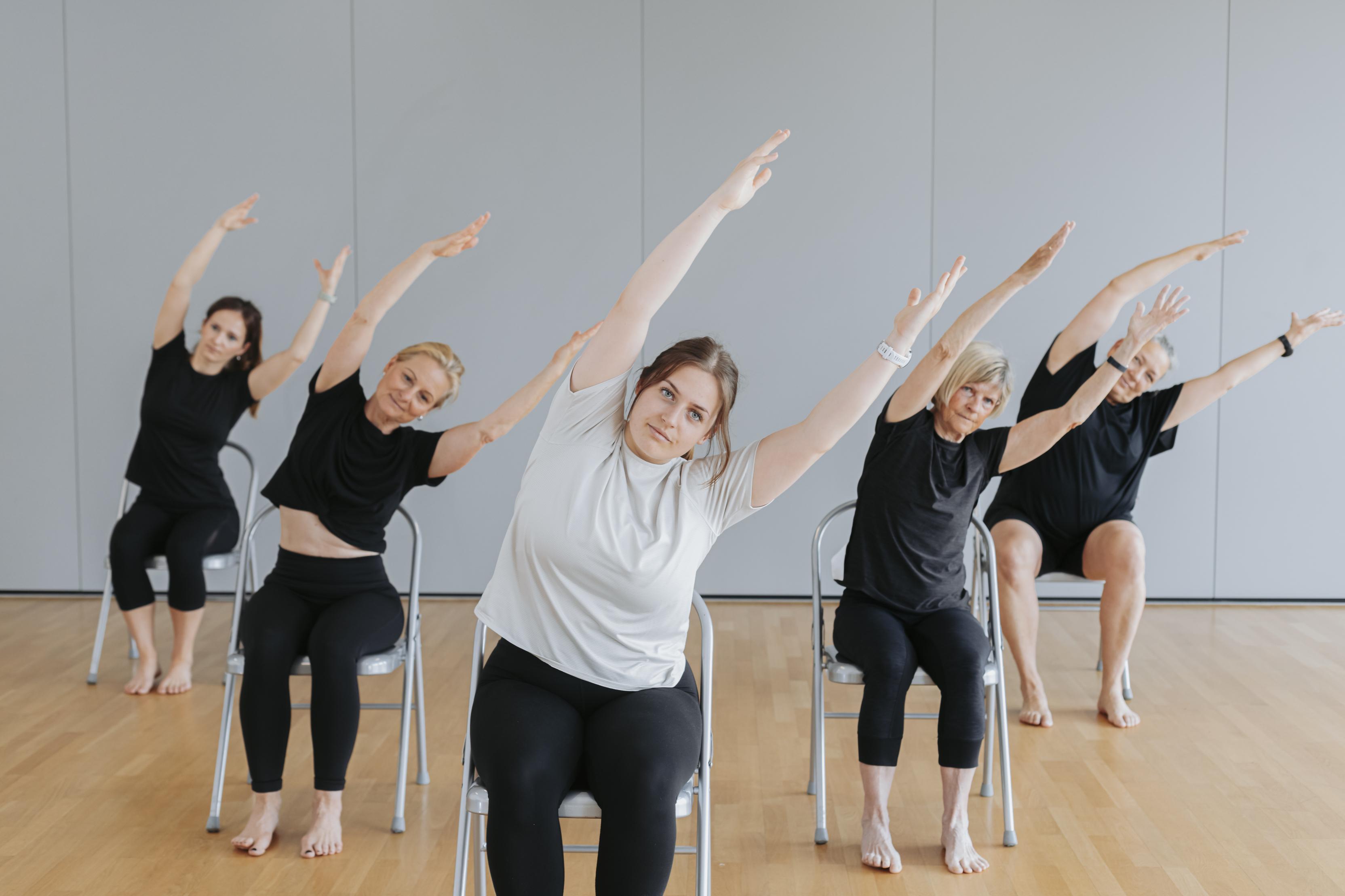 F&uuml;nf Personen sitzen in St&uuml;hlen und strecken ihre Arme in verschiedene Richtungen w&auml;hrend einer &Uuml;bung.