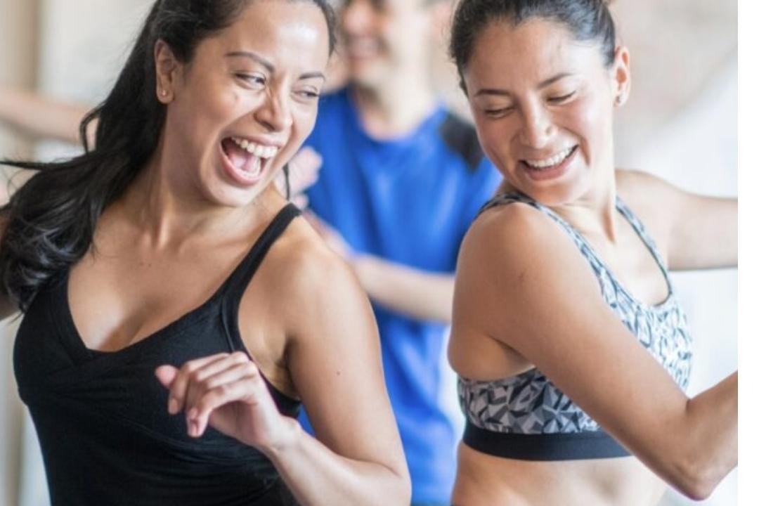 Zwei Frauen tanzen lächelnd in sportlicher Kleidung, während ein Mann im Hintergrund ebenfalls fröhlich mitmacht.