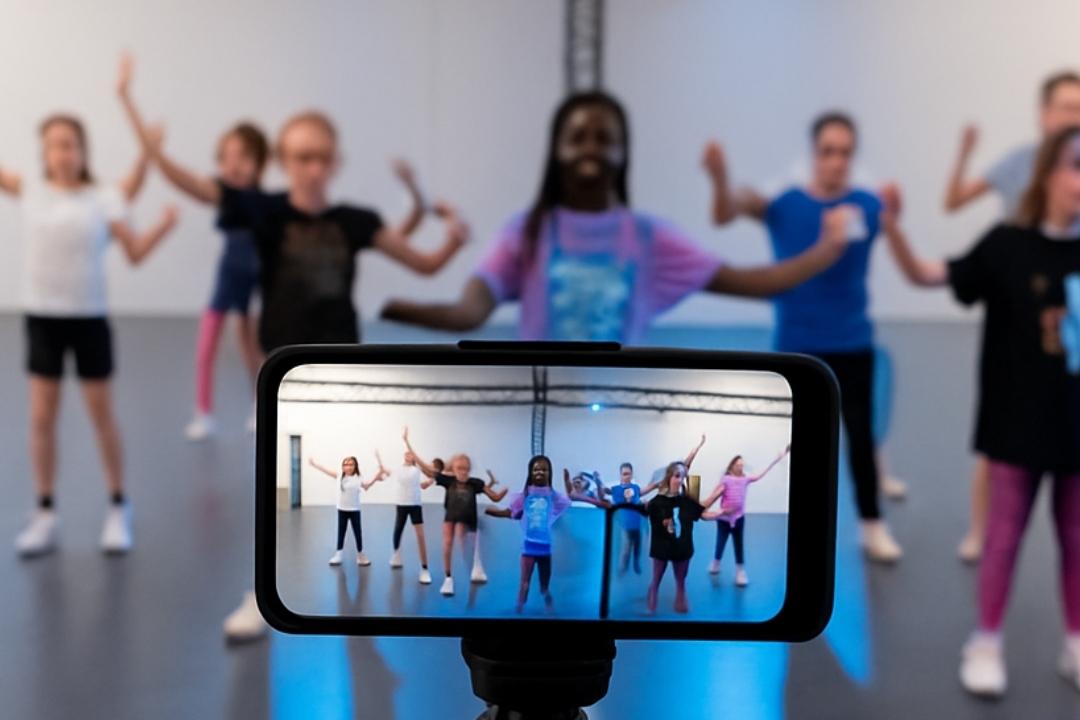 Smartphone auf einem Stativ filmt eine Tanzgruppe mit mehreren M&auml;dchen in einem lichtdurchfluteten Raum.