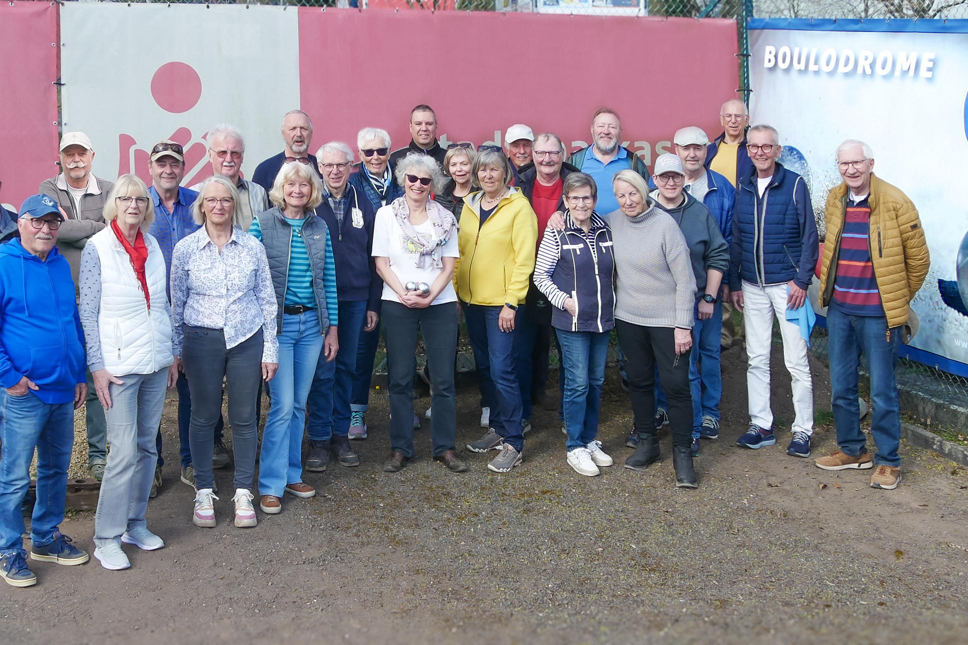 Gruppe von 25 Personen posiert drau&szlig;en vor einer Boule-Anlage, umgeben von roten und blauen Werbebanden.