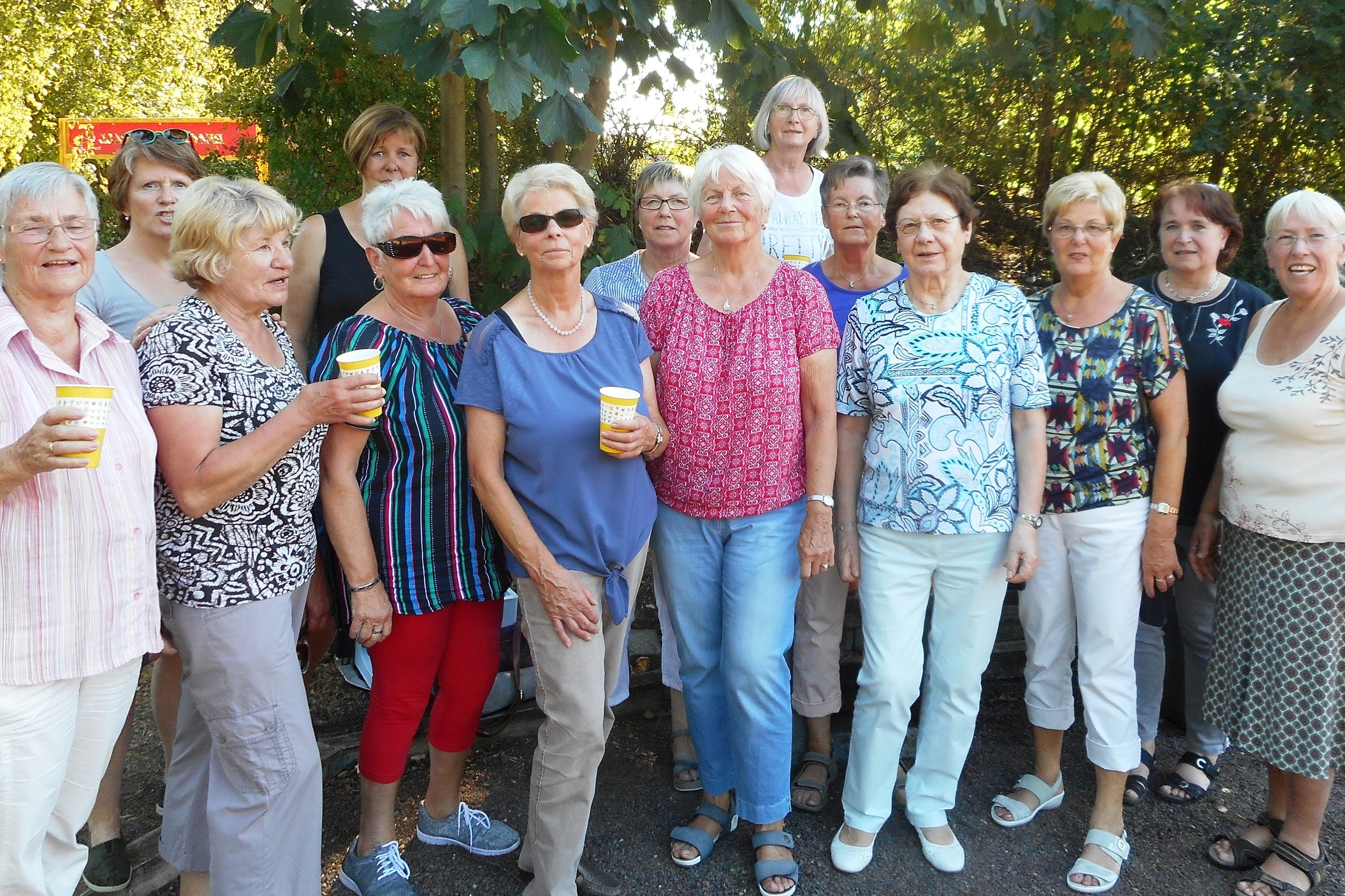 Gruppe von 15 Frauen in Freizeithaltung, l&auml;chelnd, mit Getr&auml;nken, umgeben von B&auml;umen und gr&uuml;nem Umfeld.