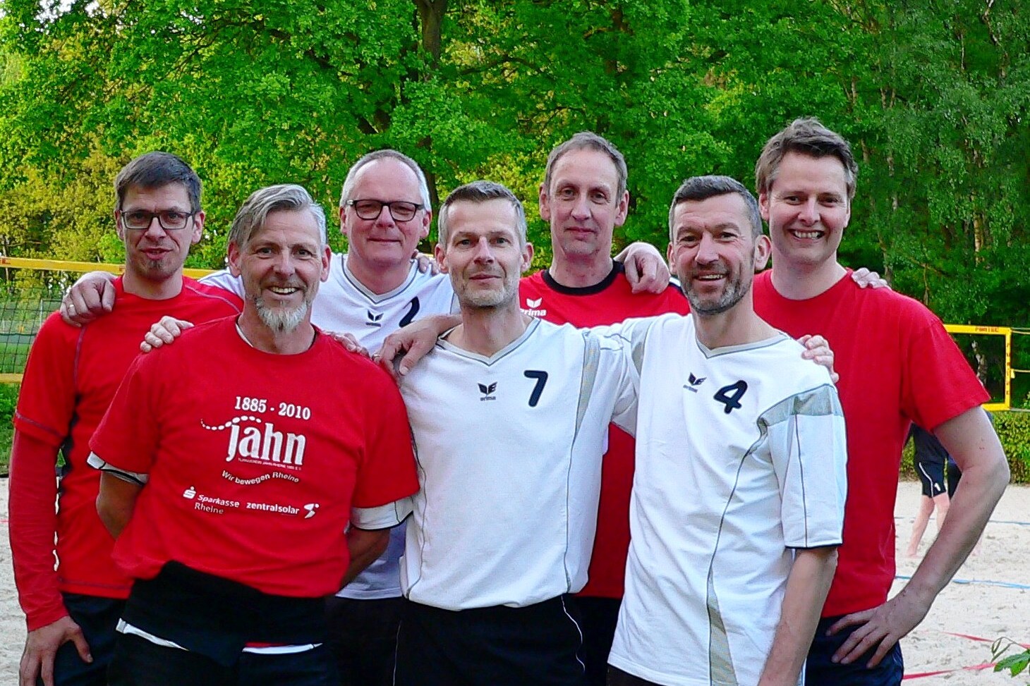 Sieben M&auml;nner in Sportkleidung posieren l&auml;chelnd auf einem Sportplatz, umgeben von B&auml;umen im Hintergrund.