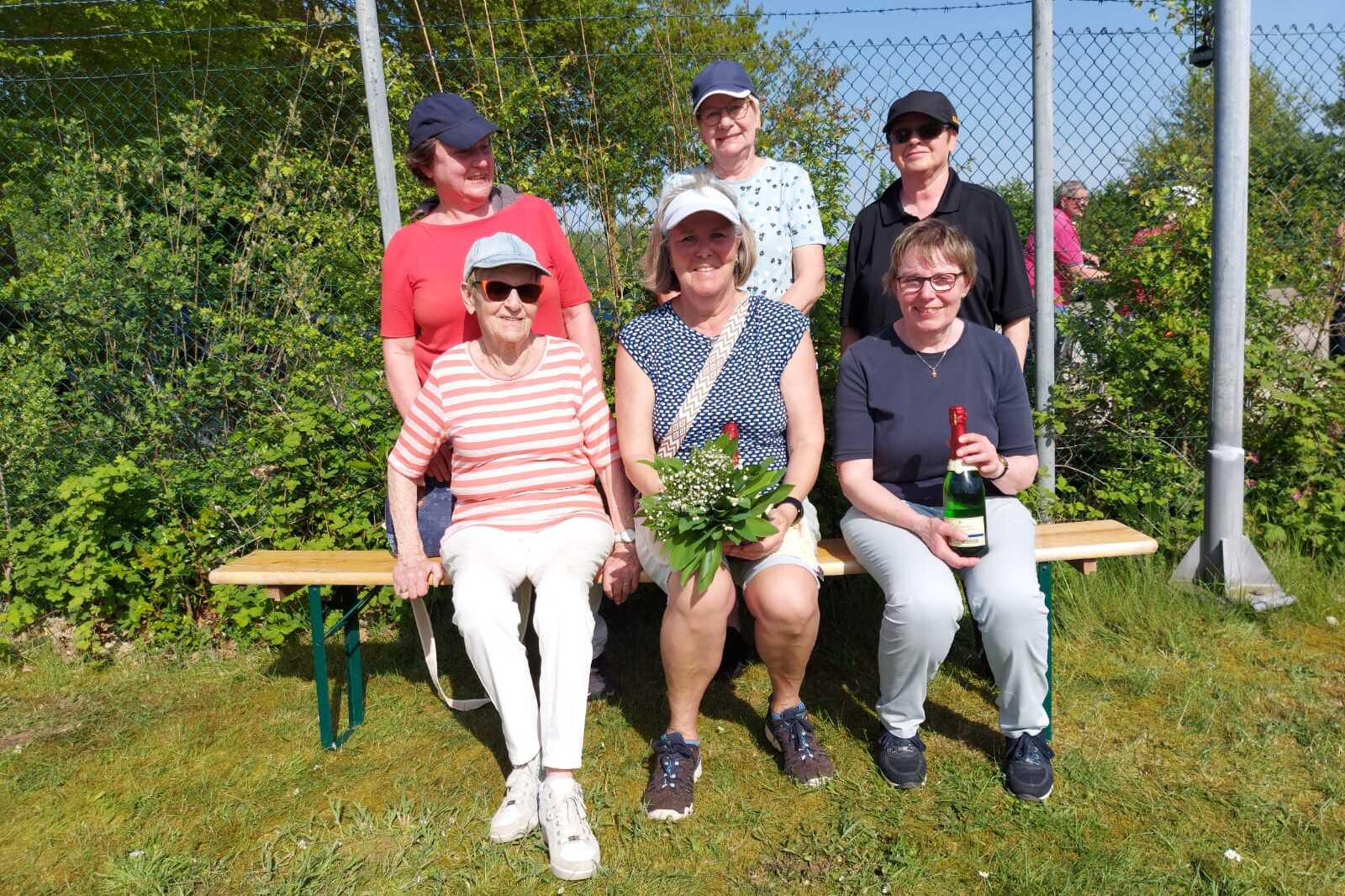 Gruppennahaufnahme von sechs Frauen auf einer Bank im Freien, umgeben von Gr&uuml;n, mit einer Pflanze und einem Sektflasche.