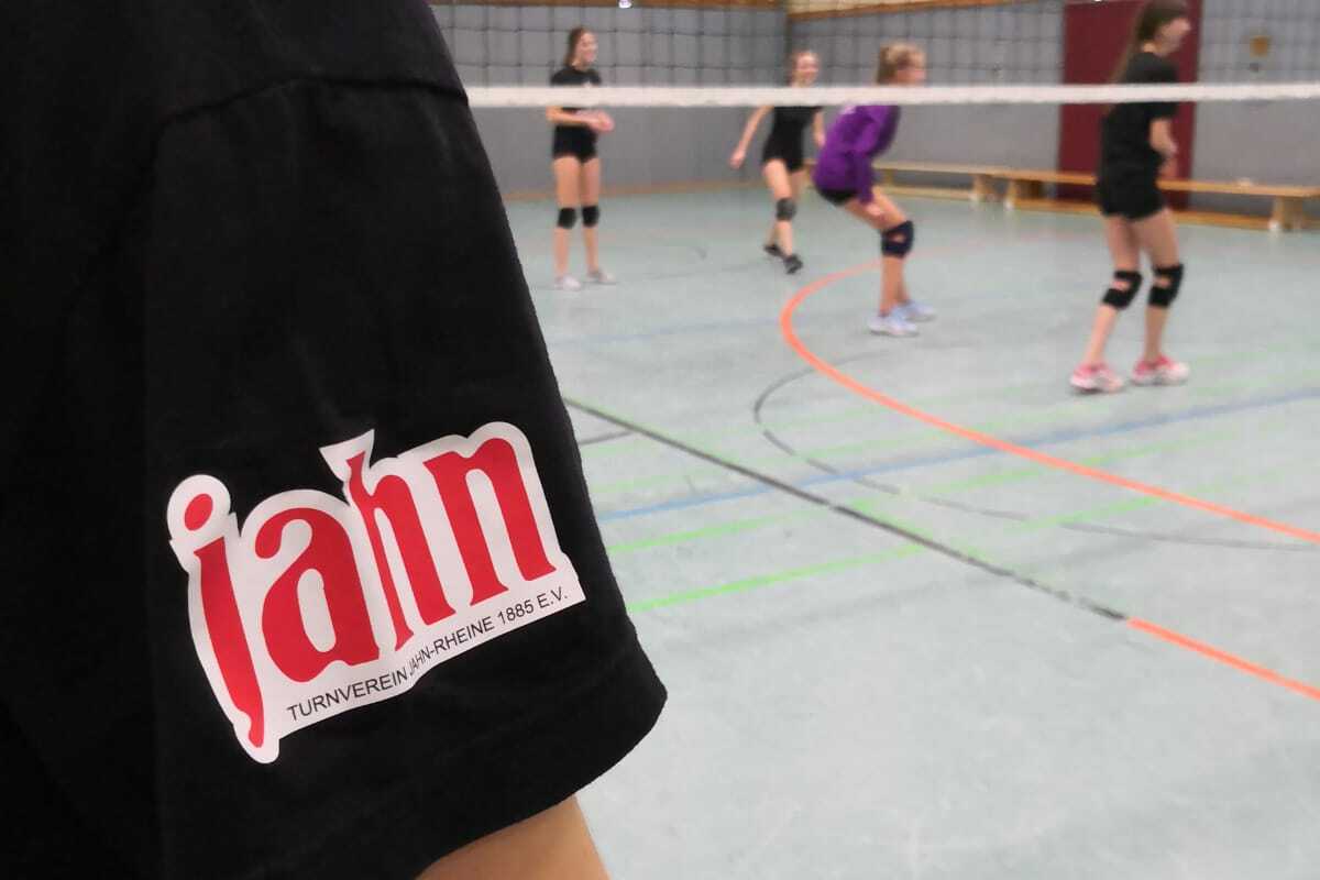 Nahaufnahme eines M&auml;dchens in einem schwarzen T-Shirt mit Vereinslogo, w&auml;hrend im Hintergrund Volleyballspielerinnen aktiv sind.