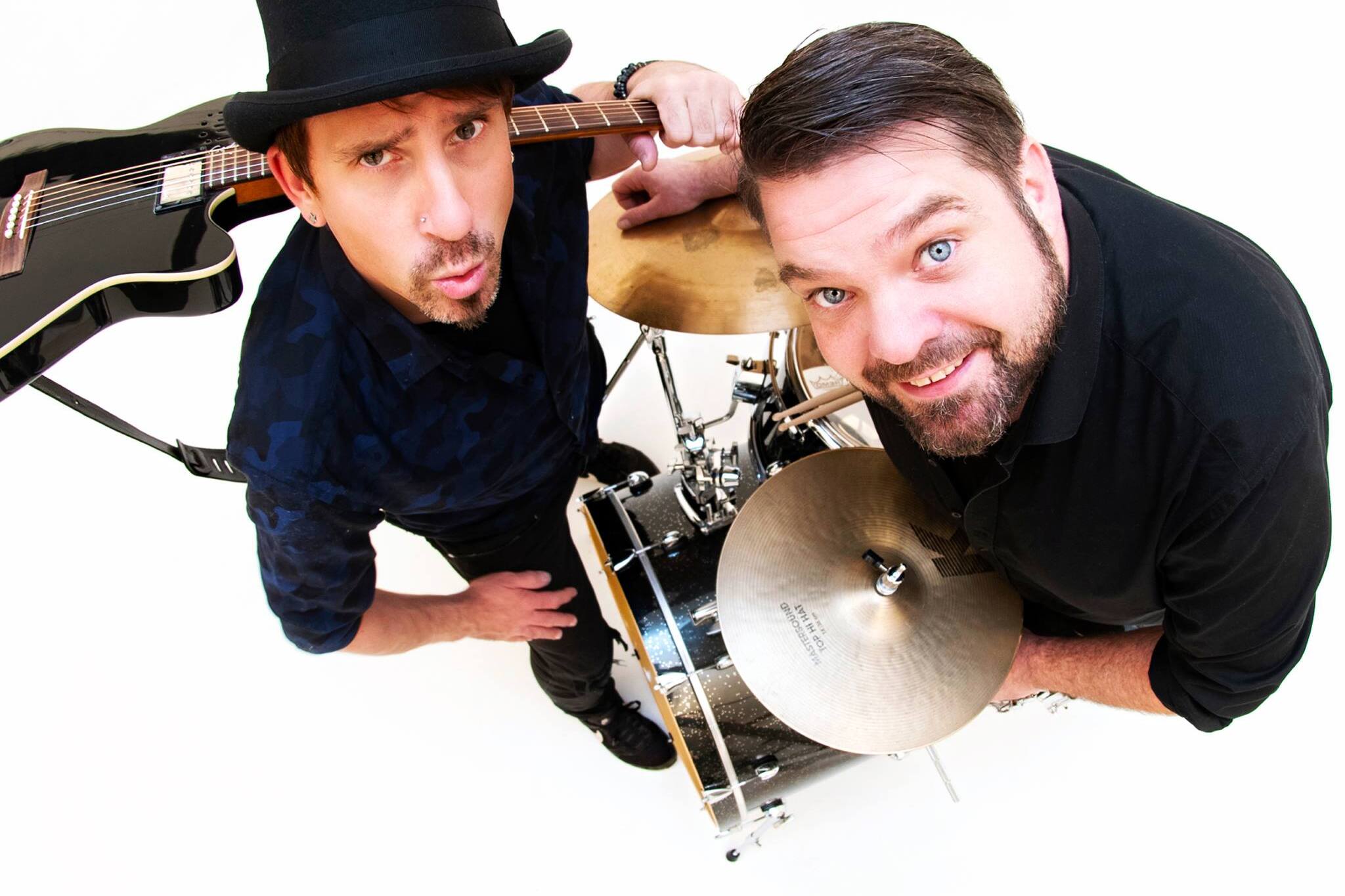 Zwei Musiker posieren: einer mit Gitarre und Hut, der andere am Schlagzeug, beide lächeln ins Kameraobjektiv.