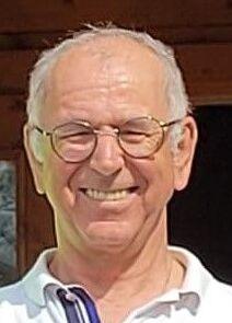 Älterer Mann mit Brille und lächelndem Gesicht, trägt ein weißes Poloshirt und steht vor einem Holzgebäude.