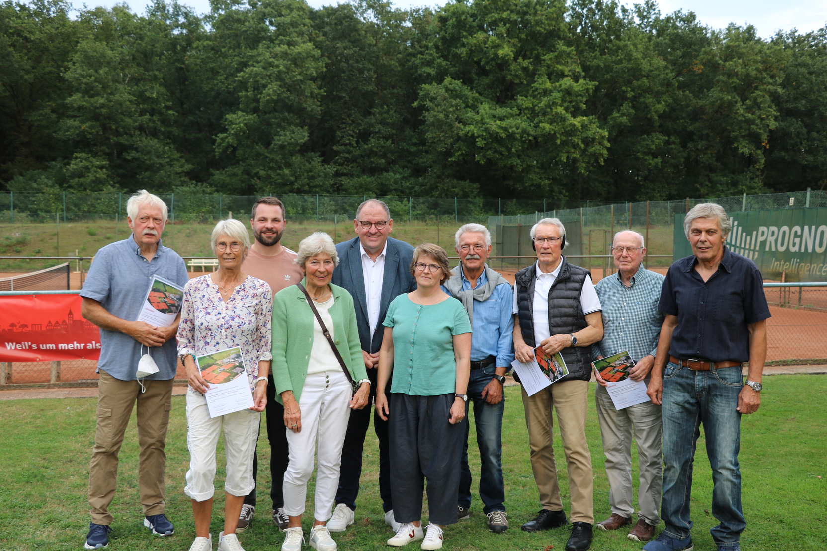 Gruppe von zehn Personen, die auf einer Tennisanlage stehen, umgeben von B&auml;umen, mit Brosch&uuml;ren in der Hand.
